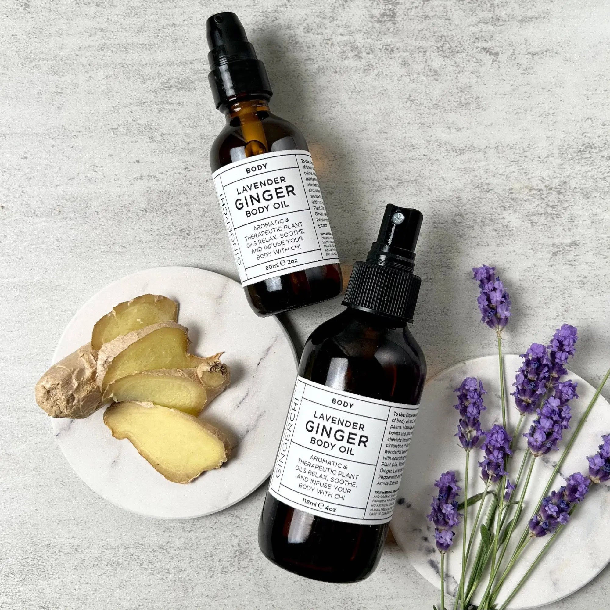 Two bottles of Lavender Ginger Body Oil with spray tops, placed next to fresh ginger roots and lavender flowers on a light background.
